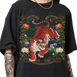 K&ouml;nnte beinhalten: Dunkelgraues T-Shirt mit einer detaillierten Illustration eines Tigers mit roten und schwarzen Streifen, umgeben von Blumen und Bl&auml;ttern. Der Tiger ist das zentrale Element, mit einem detaillierten Design. Das T-Shirt hat kurze &Auml;rmel und eine lockere Passform.