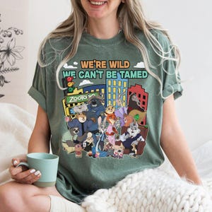 May include: Green t-shirt featuring a colorful graphic of characters from the animated movie Zootopia, with the text "WE'RE WILD WE CAN'T BE TAMED". The design includes a cityscape and various animal characters.