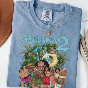 May include: A blue short-sleeve t-shirt featuring a graphic print of characters from the movie Moana 2, including Maui, Moana, and Pua. The shirt has a crew neckline and a Comfort Colors brand label on the inside collar with size S printed on it.
