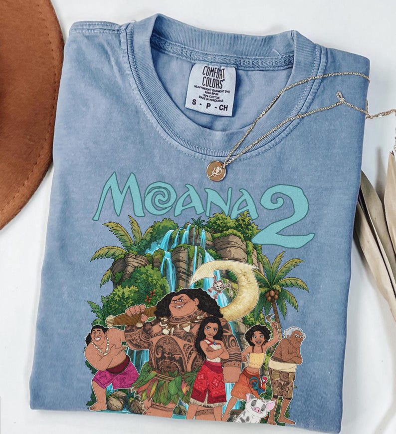 May include: A blue short-sleeve t-shirt featuring a graphic print of characters from the movie Moana 2, including Maui, Moana, and Pua. The shirt has a crew neckline and a Comfort Colors brand label on the inside collar with size S printed on it.