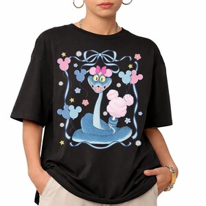 May include: Black t-shirt featuring a cartoon blue snake holding cotton candy. The snake has a pink bow and is surrounded by Mickey Mouse-shaped balloons and flowers. The design is framed by a white border with a bow at the top.