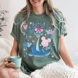 May include: Sage green t-shirt featuring a cartoon snake holding cotton candy. The snake is blue with a pink bow and Mickey Mouse-shaped balloons. The design is framed with a blue ribbon and flowers.