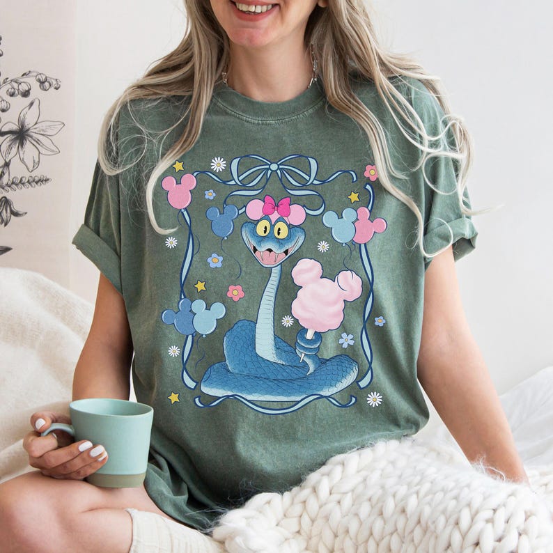 May include: Sage green t-shirt featuring a cartoon snake holding cotton candy. The snake is blue with a pink bow and Mickey Mouse-shaped balloons. The design is framed with a blue ribbon and flowers.
