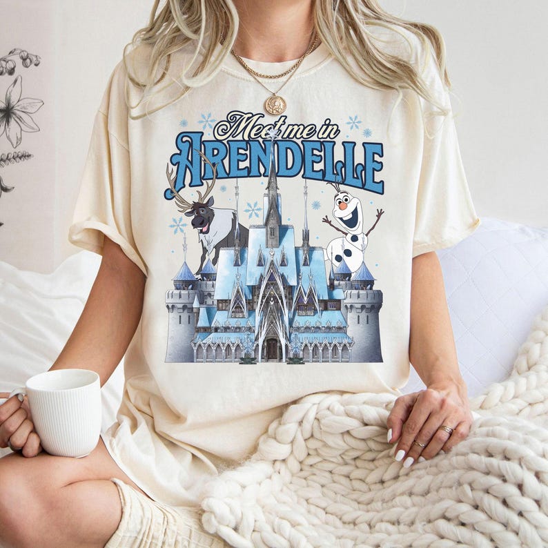 May include: Cream-colored t-shirt featuring the text "Meet me in Arendelle" above a graphic of a blue castle, a reindeer, and a snowman. The shirt has short sleeves and a relaxed fit.