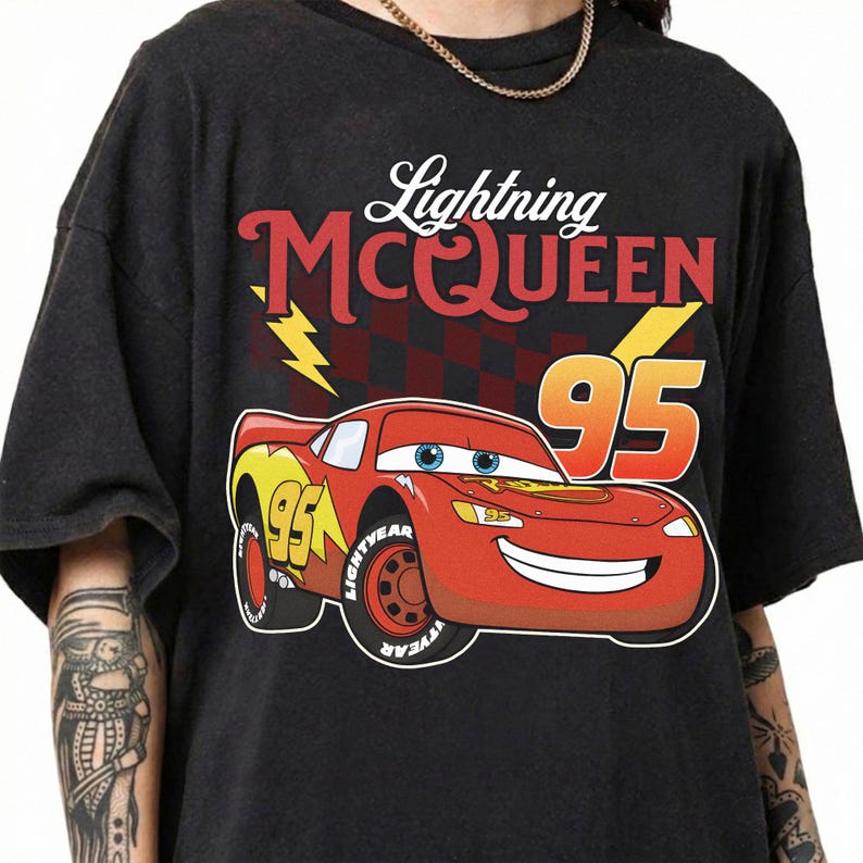 May include: A black t-shirt featuring a graphic of the red race car Lightning McQueen with the number 95, yellow lightning bolts, and a checkered pattern background. The text Lightning MCQUEEN is printed in red and white above the car graphic.