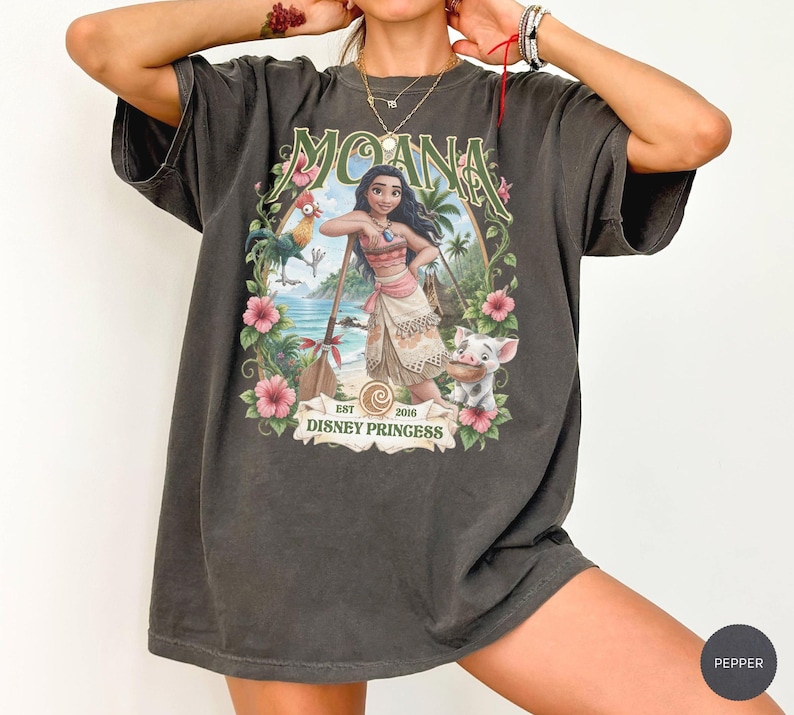 May include: A charcoal gray short-sleeve t-shirt featuring a graphic print of the character Moana, a rooster, and a piglet. The design includes tropical flowers, a wooden oar, and the text MOANA and EST 2016 DISNEY PRINGESS in a vintage style.