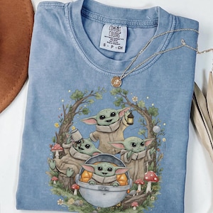 May include: A blue crew neck t-shirt featuring a graphic print of four small green creatures with large ears. One creature sits in a floating pod, while others hold a lantern, a mug, and stand among trees, mushrooms, and flowers. The label reads Comfort Colors.