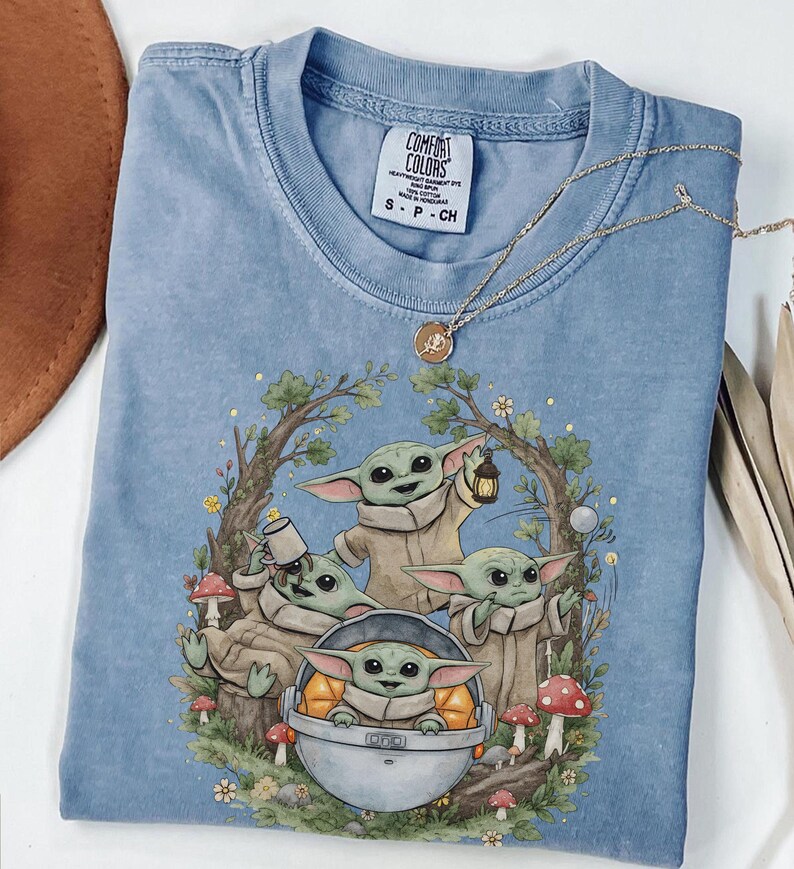 May include: A blue crew neck t-shirt featuring a graphic print of four small green creatures with large ears. One creature sits in a floating pod, while others hold a lantern, a mug, and stand among trees, mushrooms, and flowers. The label reads Comfort Colors.