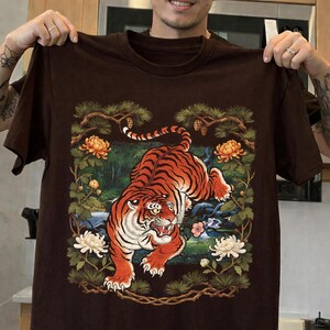 K&ouml;nnte beinhalten: Braunes T-Shirt mit einer detaillierten Illustration eines Tigers mit orangefarbenen und schwarzen Streifen, umgeben von Blumen und Bl&auml;ttern. Das Design umfasst orange und wei&szlig;e Blumen sowie Kiefernzweige.