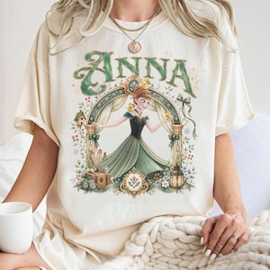 May include: A cream-colored t-shirt featuring a graphic illustration of a character named Anna in a green dress, surrounded by an ornate archway, small yellow birds, a lantern, and floral accents. The name ANNA is printed in large green letters.