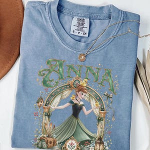 May include: A blue t-shirt featuring a graphic of a character named Anna in a green dress surrounded by yellow ducks, a birdhouse, and a lantern. The text "ANNA" is printed in large green letters above the character. A gold necklace rests on the shirt.