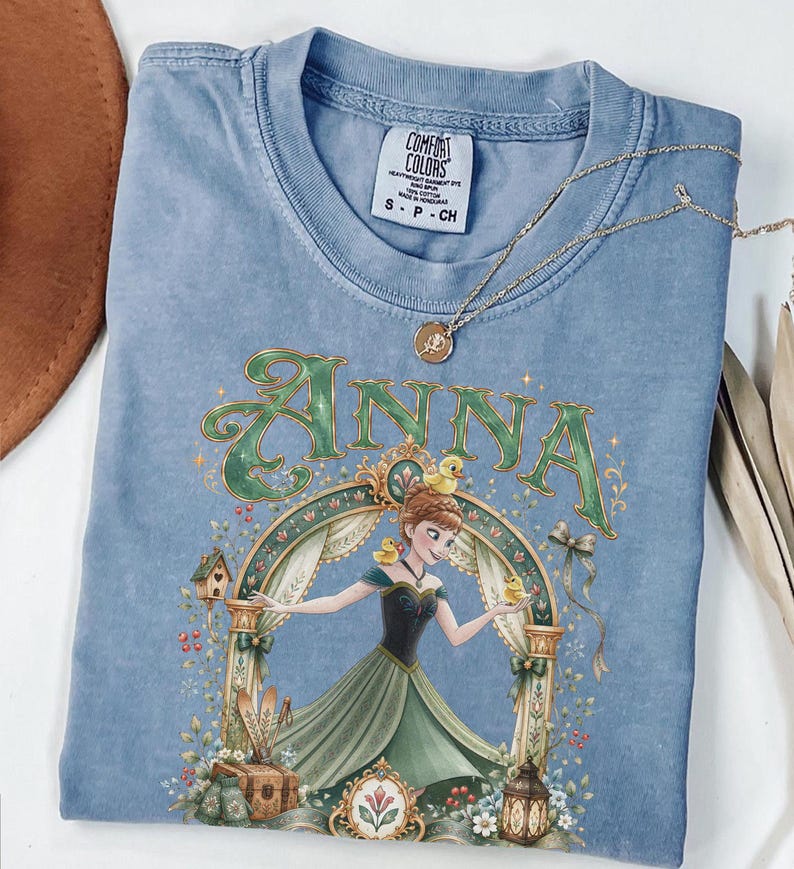 May include: A blue t-shirt featuring a graphic of a character named Anna in a green dress surrounded by yellow ducks, a birdhouse, and a lantern. The text "ANNA" is printed in large green letters above the character. A gold necklace rests on the shirt.