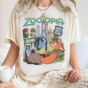 May include: A cream-colored short-sleeve t-shirt featuring a Zootopia graphic print. The design displays Judy Hopps and Nick Wilde in front of a city skyline with the text Zootopia and Bunny Best Friend. The shirt has a crew neckline and a relaxed fit.