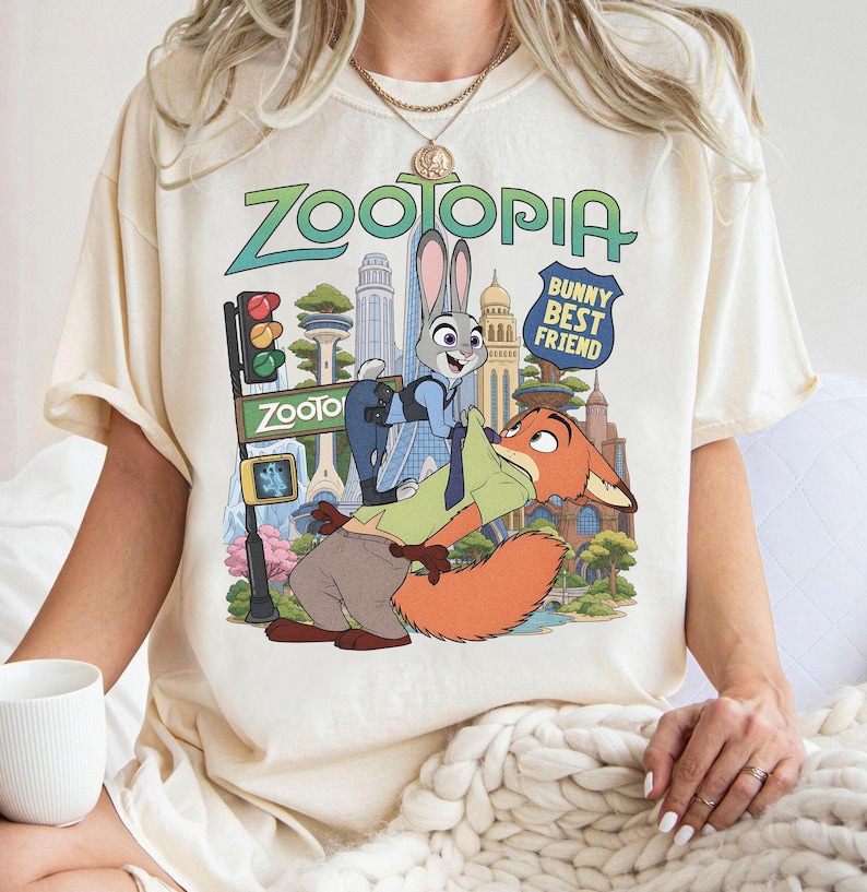 May include: A cream-colored short-sleeve t-shirt featuring a Zootopia graphic print. The design displays Judy Hopps and Nick Wilde in front of a city skyline with the text Zootopia and Bunny Best Friend. The shirt has a crew neckline and a relaxed fit.