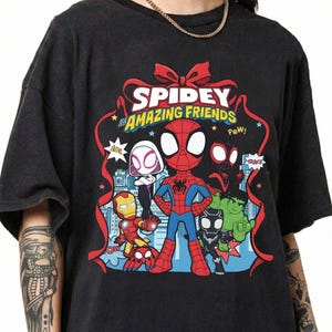 May include: A black t-shirt featuring a graphic print of Spidey and His Amazing Friends characters including Spider-Man, Ghost-Spider, Iron Man, Black Panther, and Hulk. The design is framed by a red ribbon and includes the text Spidey and His Amazing Friends.
