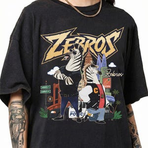 May include: A black short-sleeve t-shirt featuring a graphic print of two anthropomorphic zebras. The text "ZEBROS" appears in large yellow letters at the top. The design includes a city background, a police car, and street signs reading Hopps and Carrot.
