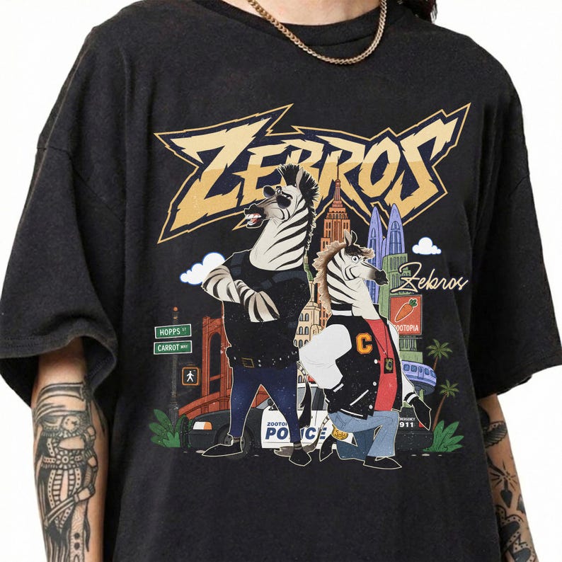 May include: A black short-sleeve t-shirt featuring a graphic print of two anthropomorphic zebras. The text "ZEBROS" appears in large yellow letters at the top. The design includes a city background, a police car, and street signs reading Hopps and Carrot.