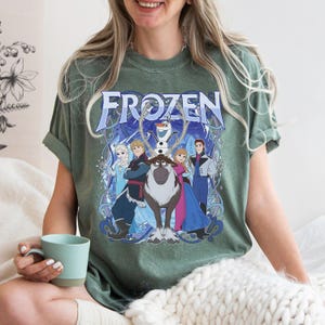 May include: A sage green short-sleeve t-shirt featuring a graphic print of the Frozen movie characters including Elsa, Anna, Kristoff, Sven, and Olaf. The word Frozen is printed in large blue letters at the top of the design with blue swirls.