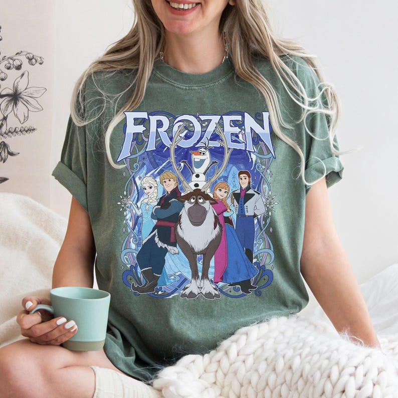 May include: A sage green short-sleeve t-shirt featuring a graphic print of the Frozen movie characters including Elsa, Anna, Kristoff, Sven, and Olaf. The word Frozen is printed in large blue letters at the top of the design with blue swirls.