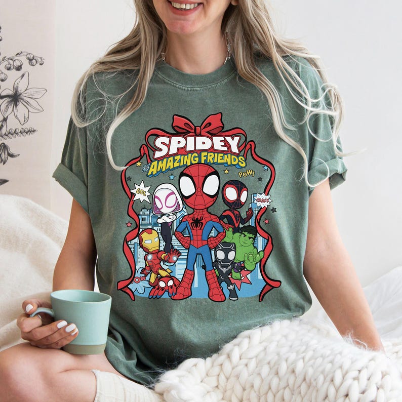May include: A sage green short-sleeve t-shirt featuring a graphic print of Spidey and His Amazing Friends characters including Spider-Man, Ghost-Spider, Iron Man, Hulk, and Black Panther, framed by a red ribbon with the text Spidey Amazing Friends.