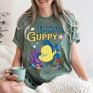 Puede incluir: Camiseta verde con la frase "I'm Not A Guppy" y una ilustraci&oacute;n de dibujos animados de un pez amarillo y azul. El dise&ntilde;o incluye coral, burbujas y una estrella de mar. La camiseta es de corte holgado.