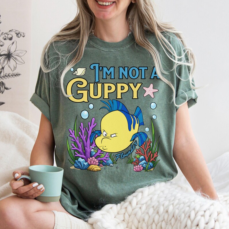 Puede incluir: Camiseta verde con la frase "I'm Not A Guppy" y una ilustraci&oacute;n de dibujos animados de un pez amarillo y azul. El dise&ntilde;o incluye coral, burbujas y una estrella de mar. La camiseta es de corte holgado.