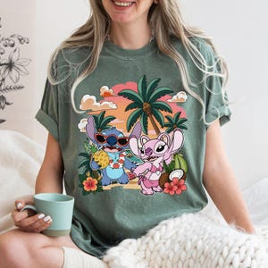May include: A sage green short-sleeve t-shirt featuring a graphic print of two blue and pink cartoon characters on a beach. The design includes palm trees, a sunset, hibiscus flowers, a pineapple, and a coconut against a soft orange sky.