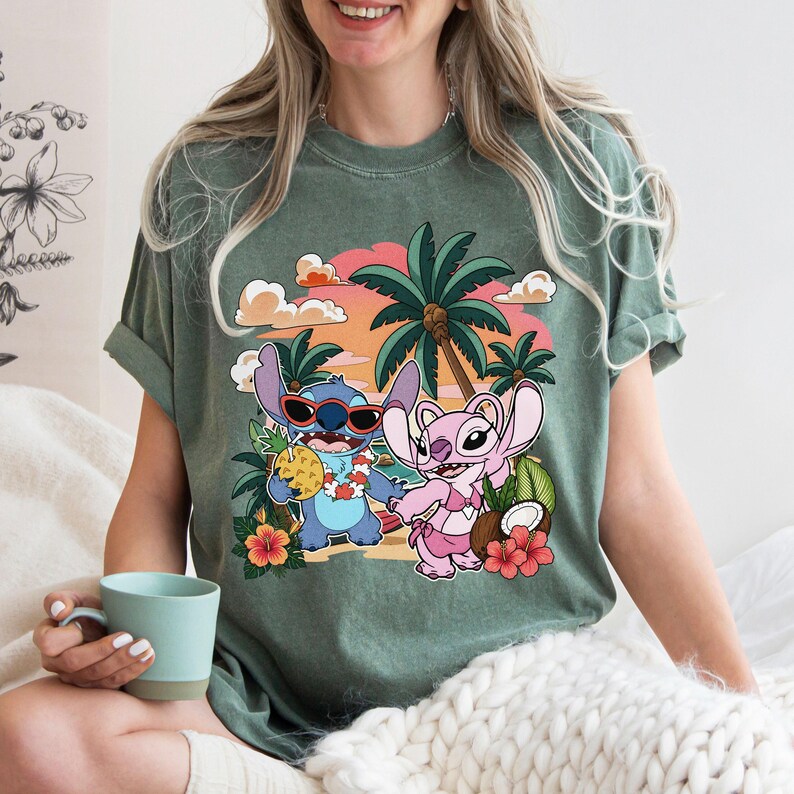 May include: A sage green short-sleeve t-shirt featuring a graphic print of two blue and pink cartoon characters on a beach. The design includes palm trees, a sunset, hibiscus flowers, a pineapple, and a coconut against a soft orange sky.