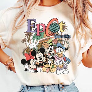 May include: Cream-colored t-shirt featuring a colorful graphic of Disney characters, including Mickey Mouse, Minnie Mouse, Goofy, Donald Duck, and Daisy Duck. The graphic includes the word "EPCOT" and "Since 1982".