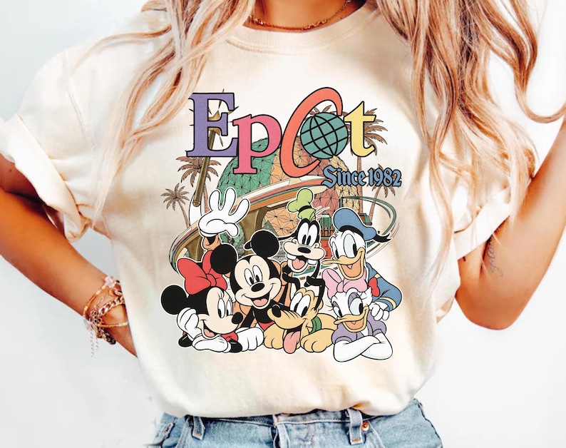 May include: Cream-colored t-shirt featuring a colorful graphic of Disney characters, including Mickey Mouse, Minnie Mouse, Goofy, Donald Duck, and Daisy Duck. The graphic includes the word "EPCOT" and "Since 1982".