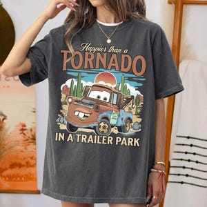 May include: Charcoal grey t-shirt with a cartoon tow truck design and the words "Happier than a Tornado in a Trailer Park." The graphic includes a desert scene with cacti and a setting sun. The shirt has short sleeves.
