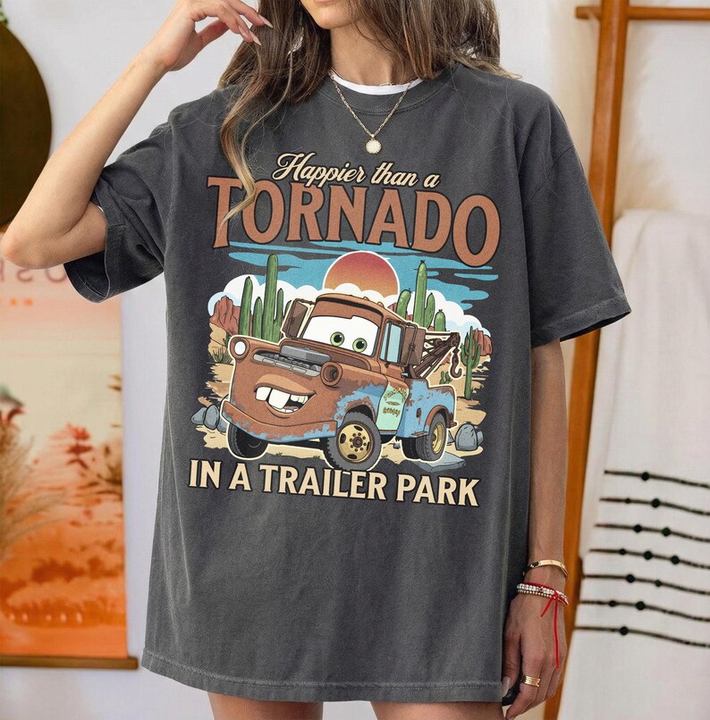 May include: Charcoal grey t-shirt with a cartoon tow truck design and the words "Happier than a Tornado in a Trailer Park." The graphic includes a desert scene with cacti and a setting sun. The shirt has short sleeves.