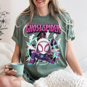 May include: Green t-shirt featuring the words "Ghost Spider" in pink and white. The shirt has a graphic of a cartoon character in a superhero pose, with a cityscape background and blue accents.