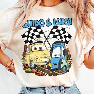 May include: Cream-colored t-shirt featuring a cartoon graphic of two cars, a yellow one and a blue one, with checkered flags. The text "Guido & Luigi" is above the cars in blue. The design is set against a brown and green background.