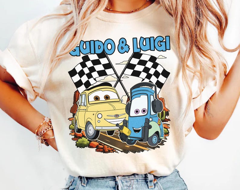 May include: Cream-colored t-shirt featuring a cartoon graphic of two cars, a yellow one and a blue one, with checkered flags. The text "Guido & Luigi" is above the cars in blue. The design is set against a brown and green background.