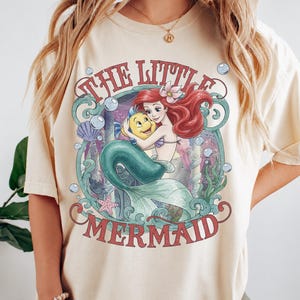 May include: A cream-colored short-sleeve t-shirt featuring a graphic print of the character Ariel from The Little Mermaid holding Flounder. The design includes red text, blue-green seaweed, bubbles, a purple seashell, and a pink starfish.