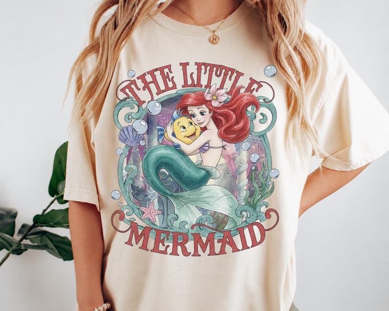 May include: A cream-colored short-sleeve t-shirt featuring a graphic print of the character Ariel from The Little Mermaid holding Flounder. The design includes red text, blue-green seaweed, bubbles, a purple seashell, and a pink starfish.