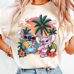 May include: A cream-colored t-shirt featuring a graphic print of two blue and pink cartoon characters on a beach. The blue character wears red sunglasses and holds a pineapple drink, while the pink character wears a bikini. Palm trees and flowers surround them.
