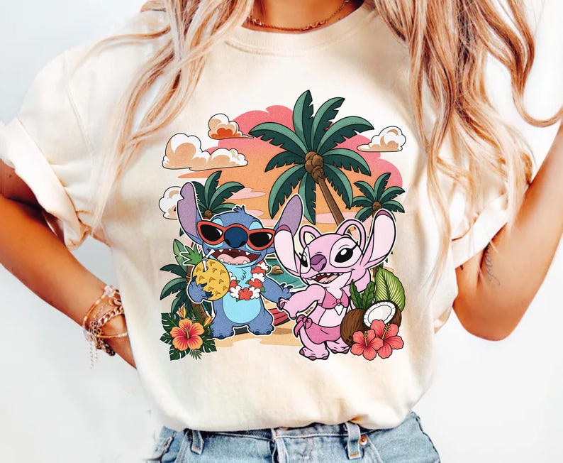 May include: A cream-colored t-shirt featuring a graphic print of two blue and pink cartoon characters on a beach. The blue character wears red sunglasses and holds a pineapple drink, while the pink character wears a bikini. Palm trees and flowers surround them.