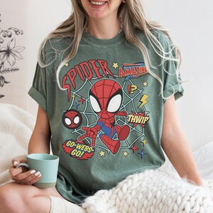 May include: A sage green short-sleeve t-shirt featuring a graphic of a cartoon Spider-Man character in a web. The design includes red text reading "SPIDER", "AMAZING", "THWIP", and "GO-WEBS GO!" along with yellow stars and lightning bolt accents.