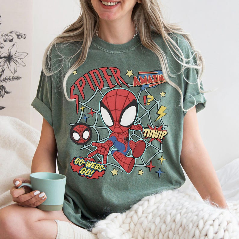 May include: A sage green short-sleeve t-shirt featuring a graphic of a cartoon Spider-Man character in a web. The design includes red text reading "SPIDER", "AMAZING", "THWIP", and "GO-WEBS GO!" along with yellow stars and lightning bolt accents.