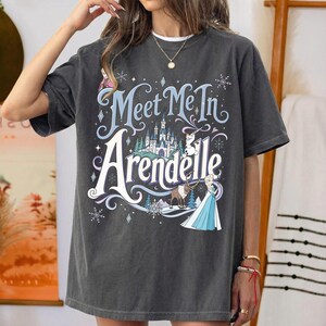K&ouml;nnte beinhalten: Ein anthrazitfarbenes Kurzarm-T-Shirt mit einem Grafikdruck und dem Text "Meet Me In Arendelle". Das Design zeigt ein Schloss, einen Schneemann, ein Rentier und eine weibliche Figur in einem blauen Kleid, umgeben von Schneeflockenmotiven.