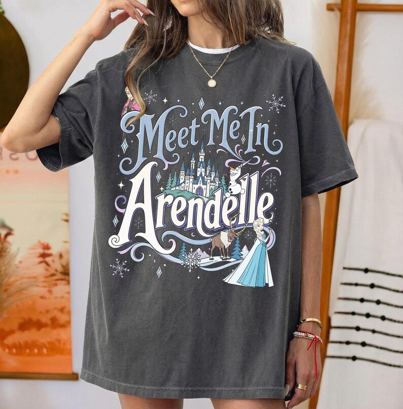 K&ouml;nnte beinhalten: Ein anthrazitfarbenes Kurzarm-T-Shirt mit einem Grafikdruck und dem Text "Meet Me In Arendelle". Das Design zeigt ein Schloss, einen Schneemann, ein Rentier und eine weibliche Figur in einem blauen Kleid, umgeben von Schneeflockenmotiven.