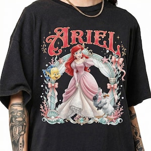May include: A black short-sleeve t-shirt featuring a graphic print of Ariel in a pink gown, surrounded by Flounder, Scuttle, and an ornate archway. The text ARIEL is printed in large red letters above the character illustration.