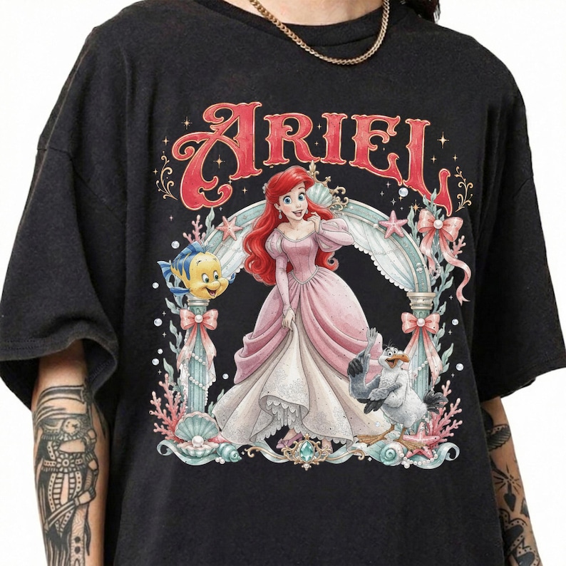 May include: A black short-sleeve t-shirt featuring a graphic print of Ariel in a pink gown, surrounded by Flounder, Scuttle, and an ornate archway. The text ARIEL is printed in large red letters above the character illustration.