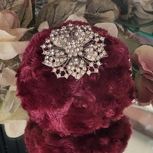 May include: A burgundy, plush, round object with a large, ornate, silver-toned brooch. The brooch is encrusted with clear, sparkling rhinestones, arranged in a floral design. The object's texture is soft and fuzzy, with a slight sheen.