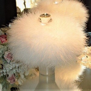 May include: A vintage powder puff with a fluffy, light pink exterior. The puff is topped with a clear, decorative jewel. The puff sits on a clear, cylindrical base, suggesting a vanity or boudoir accessory.