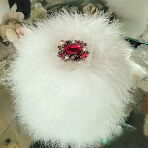 May include: A white, fluffy powder puff with a large, ornate jewel at its center. The jewel features a rectangular, red gemstone surrounded by smaller red, brown, and pearl-colored stones, set in a silver-toned metal.