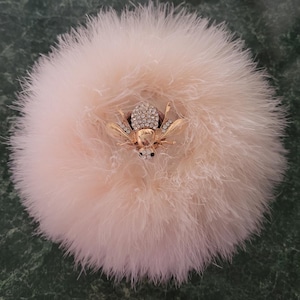 May include: A fluffy, light pink powder puff with a gold-colored bee brooch. The bee is adorned with sparkling rhinestones. The puff is round and has a soft, feathery texture.