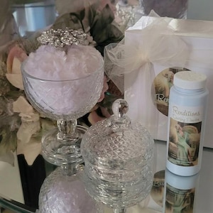 May include: A clear glass goblet filled with white fluff, a decorative jar with a lid, and a light blue bottle of Renditions Dusting Powder are displayed on a reflective surface. A sparkling silver-toned ornament sits atop the goblet.